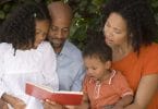 Família de afrodescendentes composta por pai, mãe, filha e filho lendo um livro no jardim.