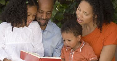 Família de afrodescendentes composta por pai, mãe, filha e filho lendo um livro no jardim.