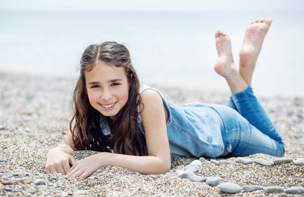 Menina deitada na areia de uma praia com cabelos escuros, bula azul e calça jeans;