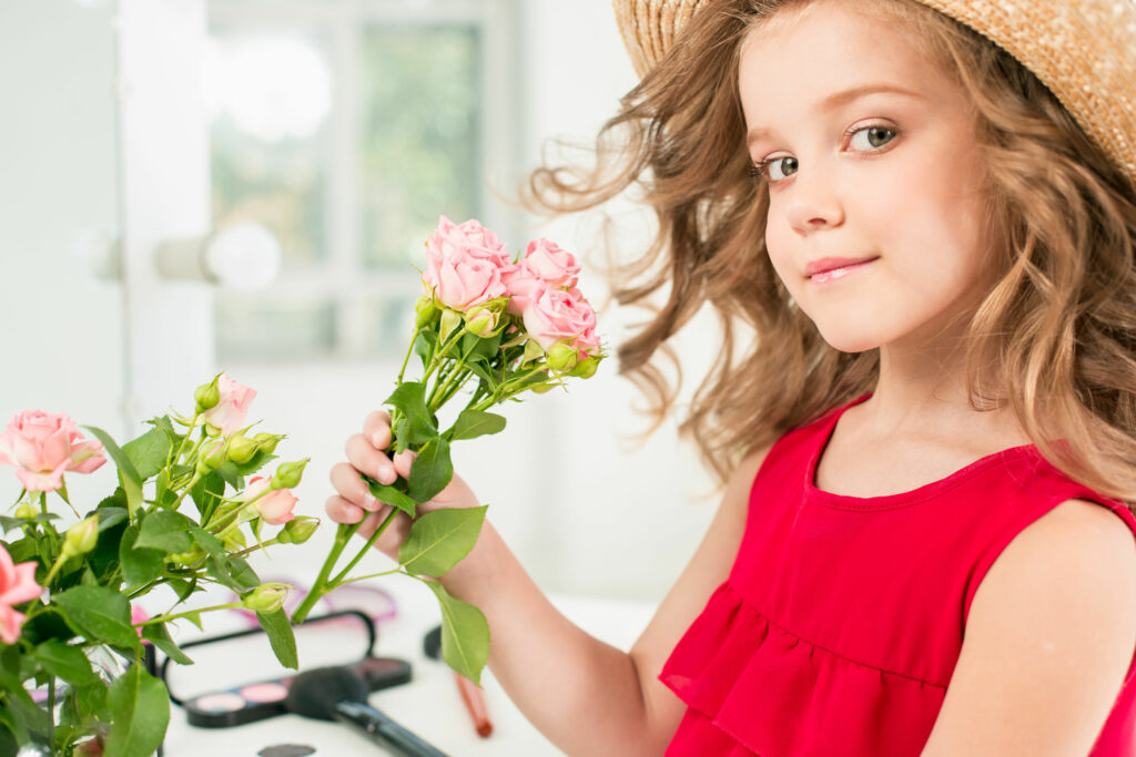 Garotinha segura rosas, ela está com vestido vermelho e com um chapéu bege.