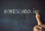 Criança escrevendo em quadro negro com giz a palavra "homeschooling" que significa Educação Domiciliar.