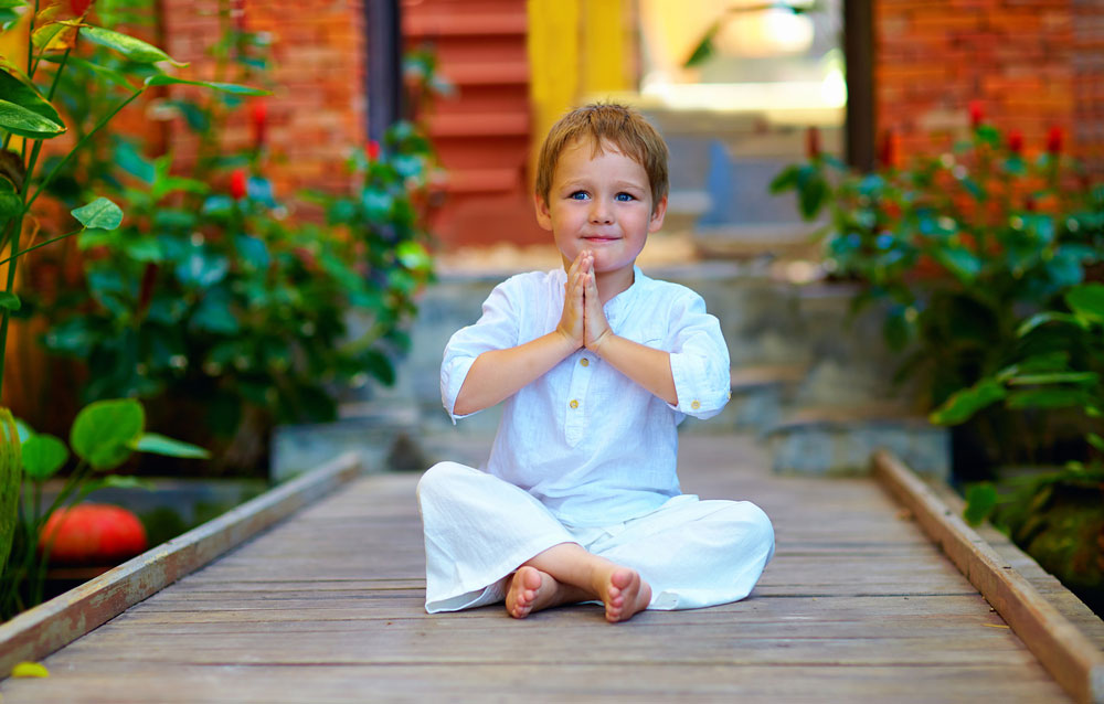 Menino de aproximadamente oito anos sentaod em posição meditativa com as pernas cruzadas e as mãos juntas sentado em chão d emadeira vestindo roupas brancas.