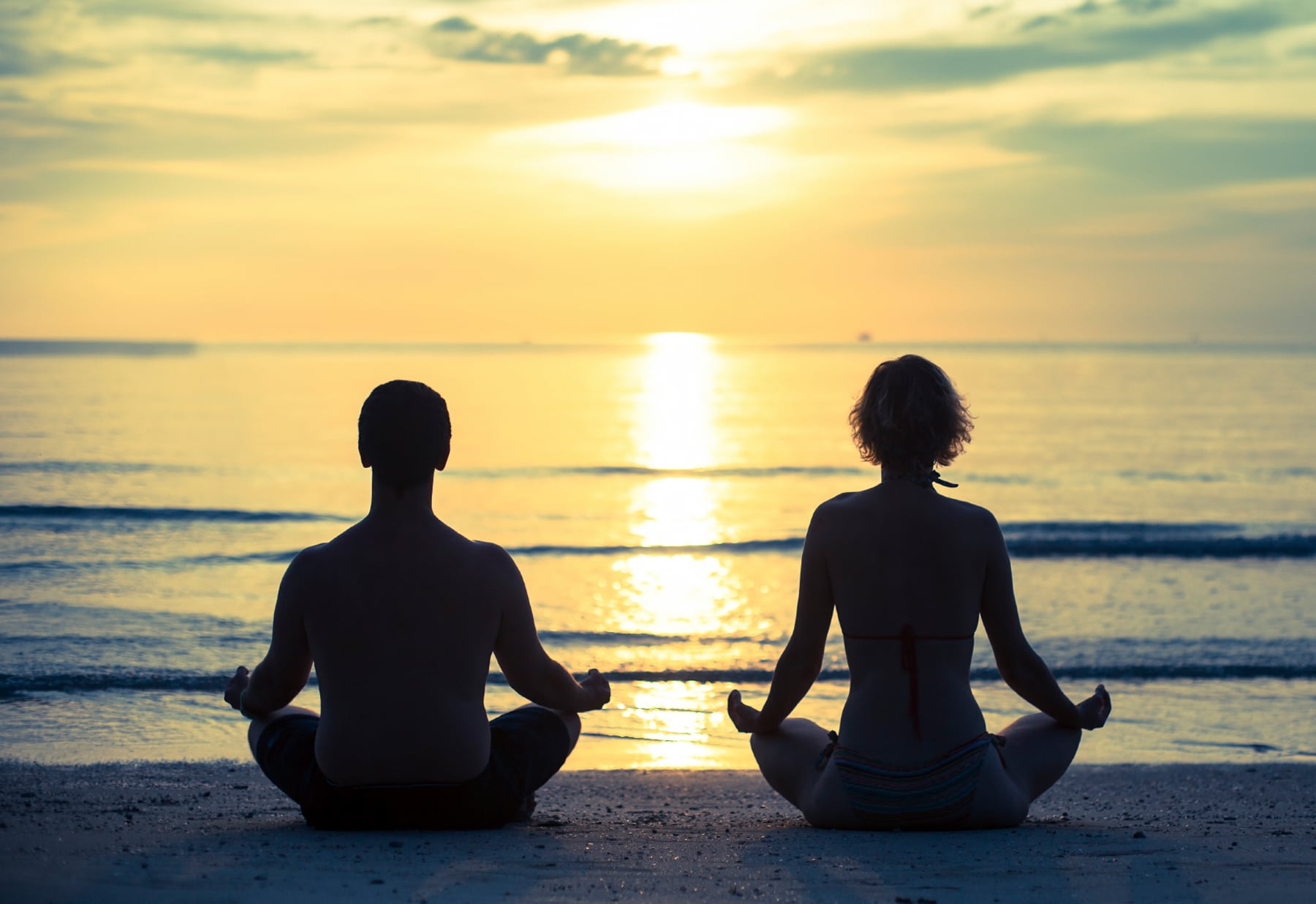 Duas pessoas sentadas de frente par ao mar meditam em posição de lotus.