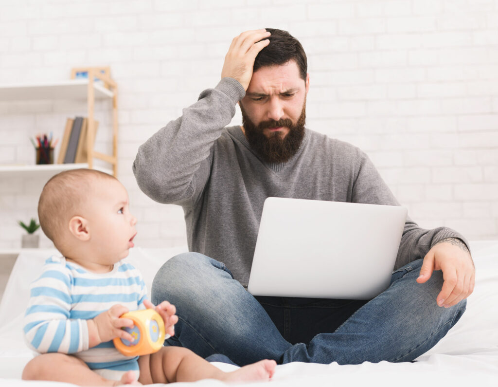 Homem barbudo sentado no chão, mexendo em um notebook, com cara de preocupado, ao lado de um bebê.