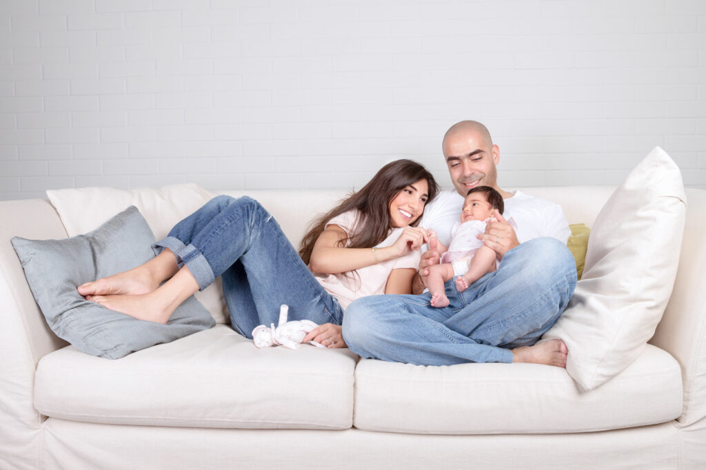 Homem e mulher brancos, sentados em um sofá branco, no meio de uma sala branca, ondem o homem segura um bebê.