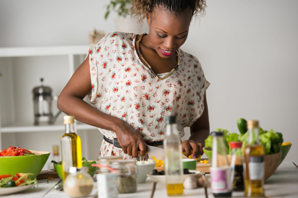 Mulher negra jovem e sorridente em uma cozinha branca cortando vegetais para fazer uma salada, cercada de utensílios e outras comidas e temperos.