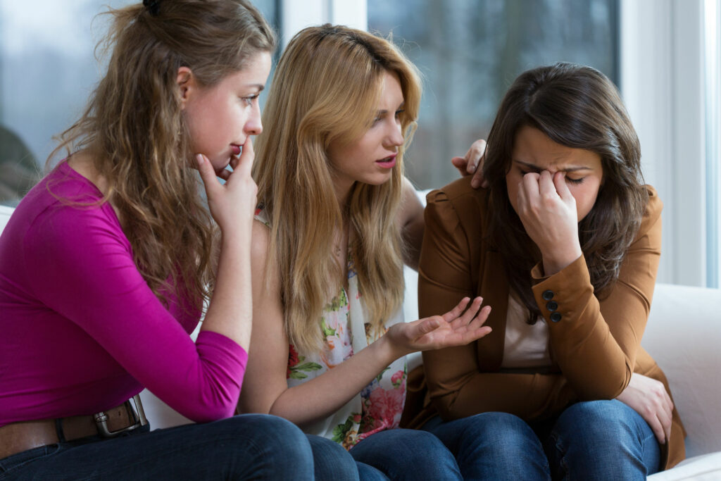 Três mulheres sentadas em um sofá, com uma delas chorando e as outras duas tentando consolá-la. 