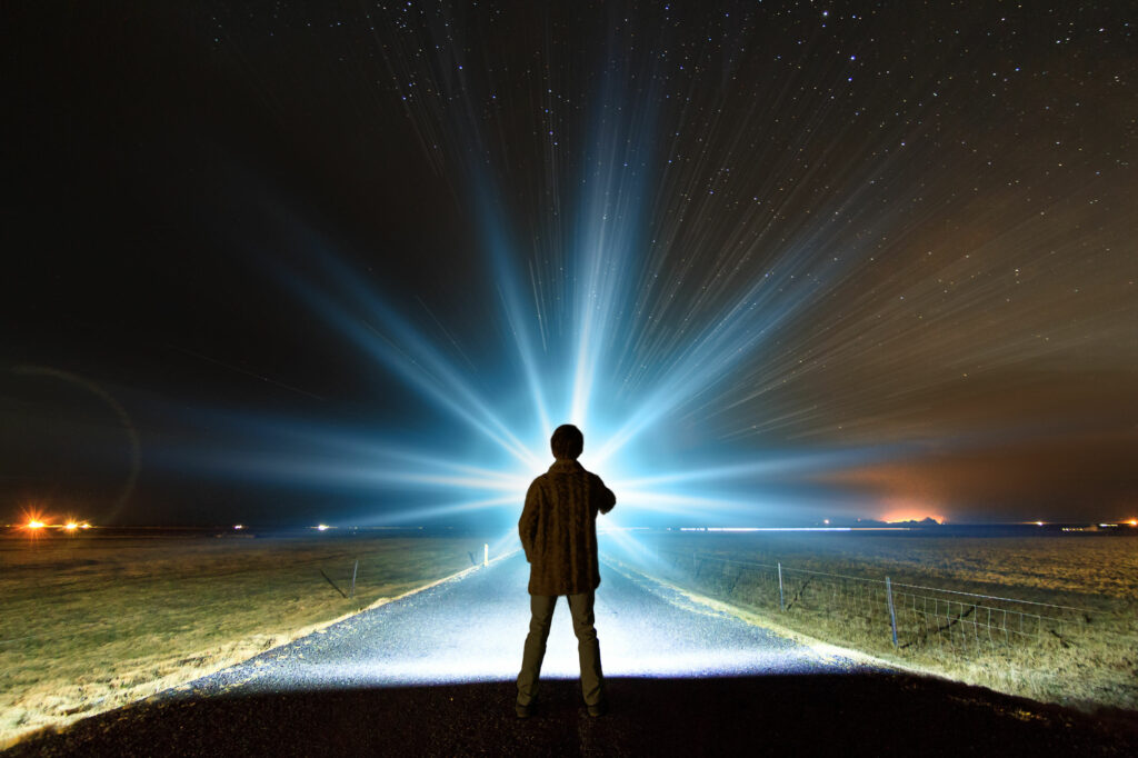 Silhueta de homem paro em uma estrada rural, olhando em direção à uma luz brilhante no horizonte noturno.