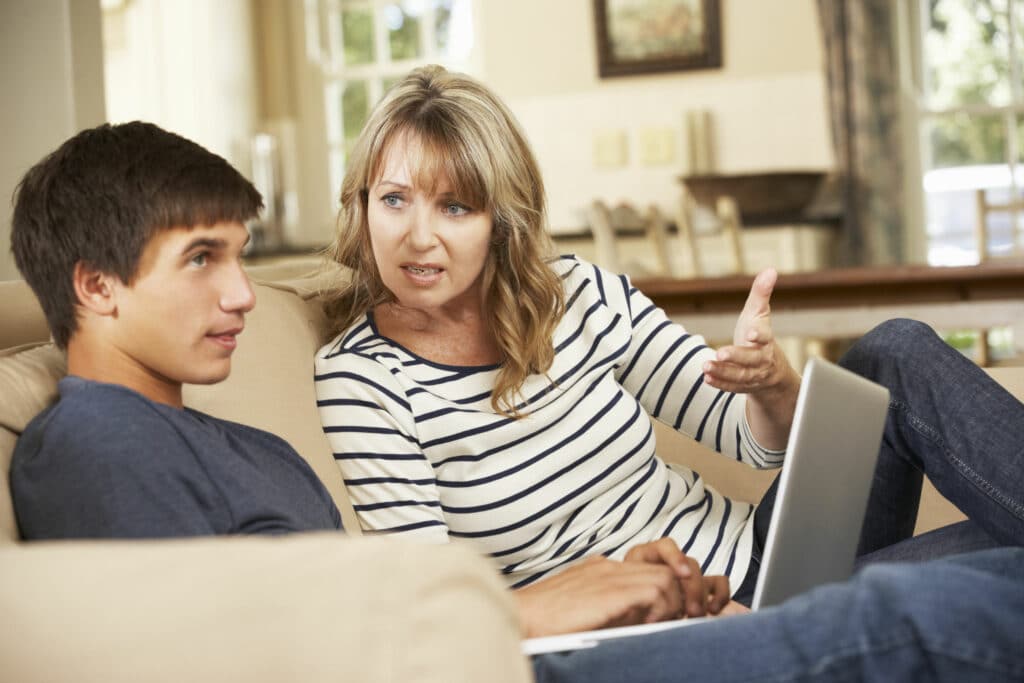 mãe e filho sentados em um sofá da sala. O menino está mexendo em um notebook e ignorando sua mãe, que está brigando com ele.