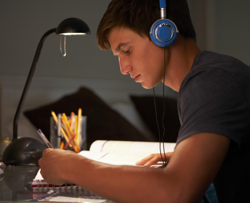 Menino branco usando fones de ouvi azuis escrevendo em um caderno com um livro aberto do lado, como se estivesse estudando.