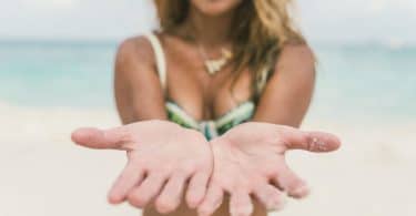 Mulher na praia com as mãos estendidas para a câmera. Ela tem o cabelo loiro e usa um biquíni verde.