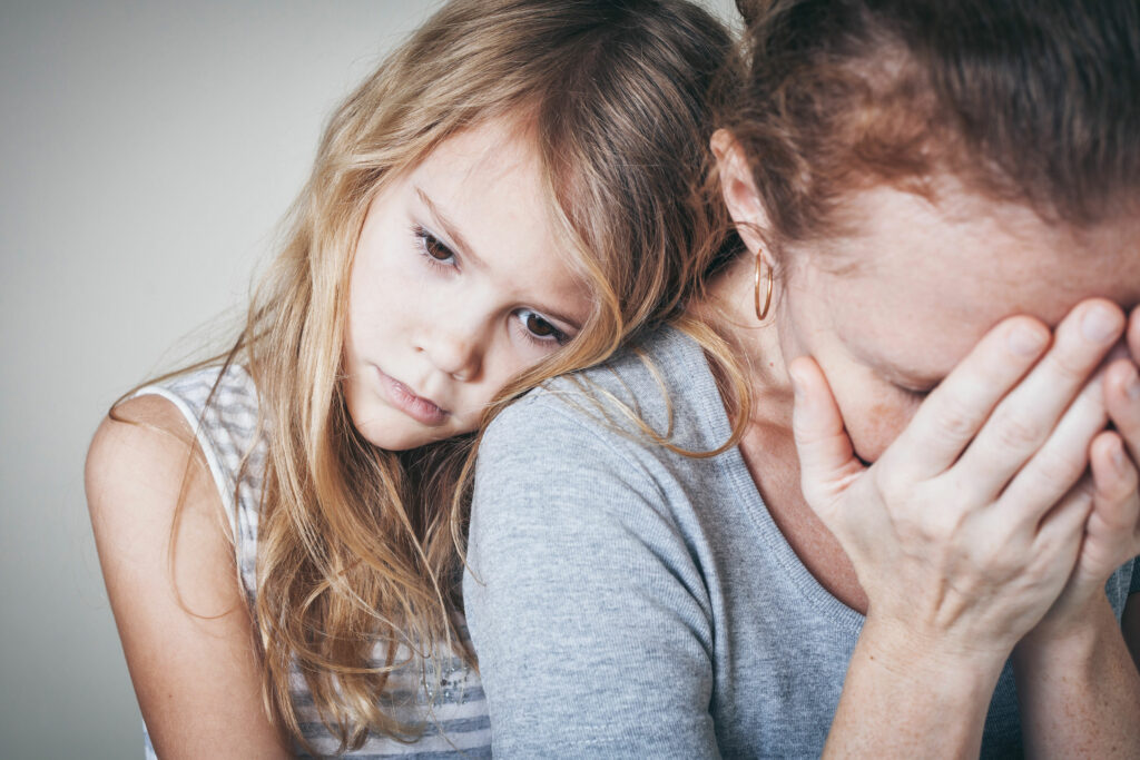 Menina criança branca, com expressão preocupada e triste, apoiando sua cabeça no ombro de sua mãe.