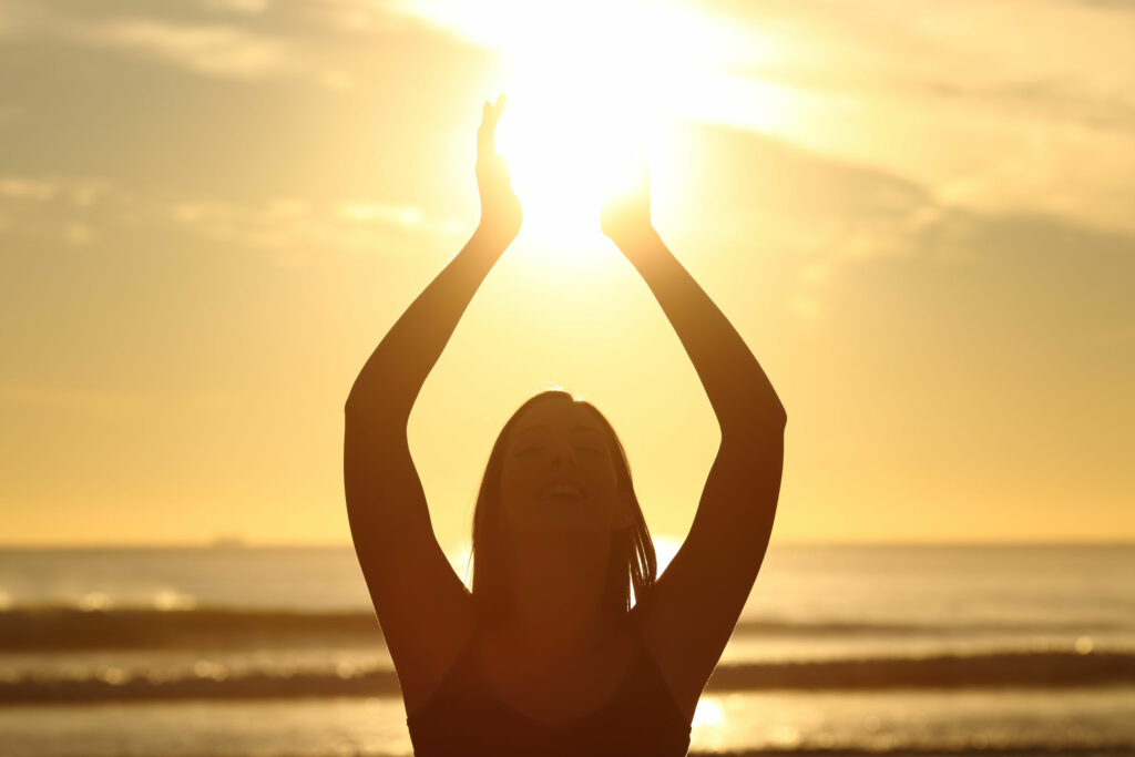 Mulher feliz na praia com os baraços para cima, tentando alcançar uma luz no céu.