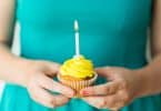 Mulher segura cupcake de cobertura amarela com uma vela acesa.