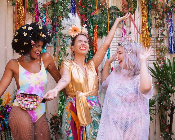 Três garotas pulando carnaval fantasiadas e cheias de glitter. Uma negra e duas brancas.