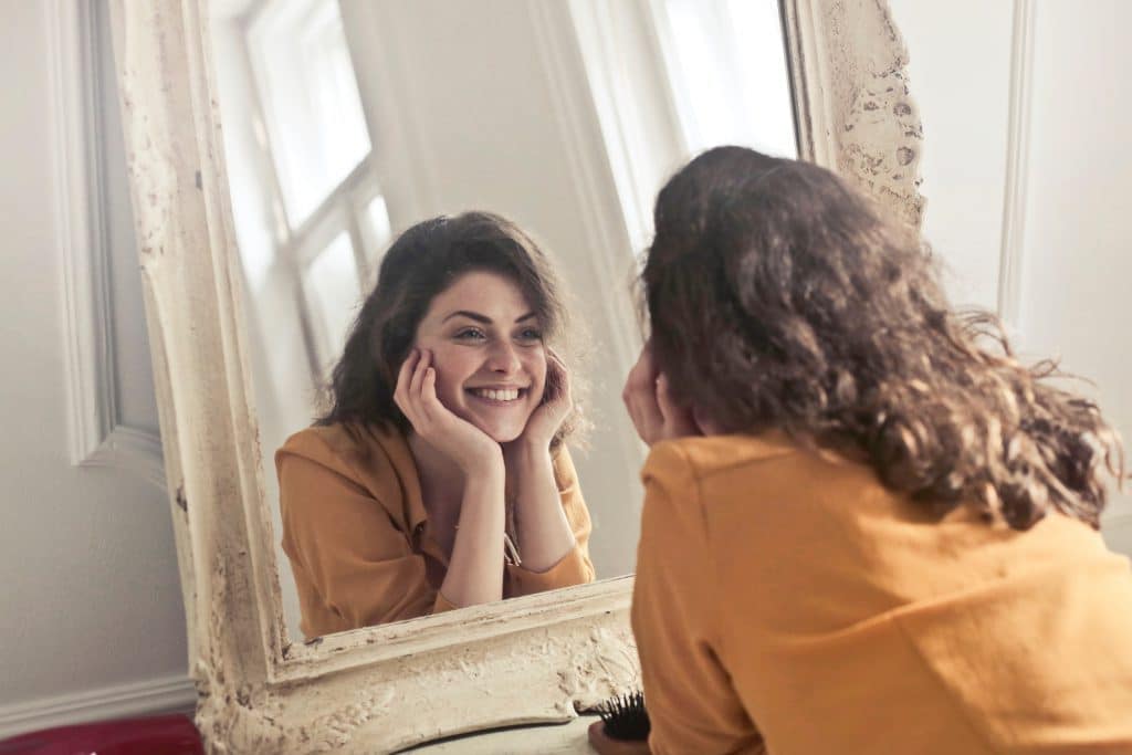Mulher sorrindo para o próprio reflexo no espelho. Usa blusa amarela e tem cabelo castanho.