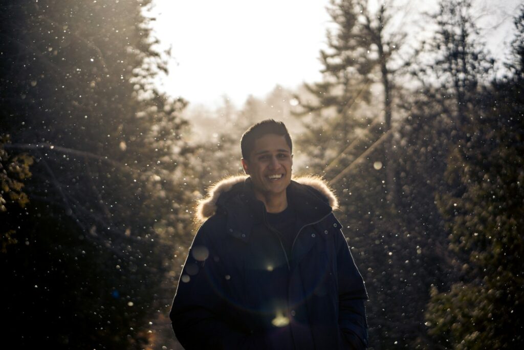 Homem sorrindo. Ele está em uma floresta iluminada pelo sol ao fim de tarde.