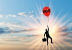 Homem carregando mala e usando uma coroa enquanto segura um balão vermelho com a a palavra "ego" desenhada.