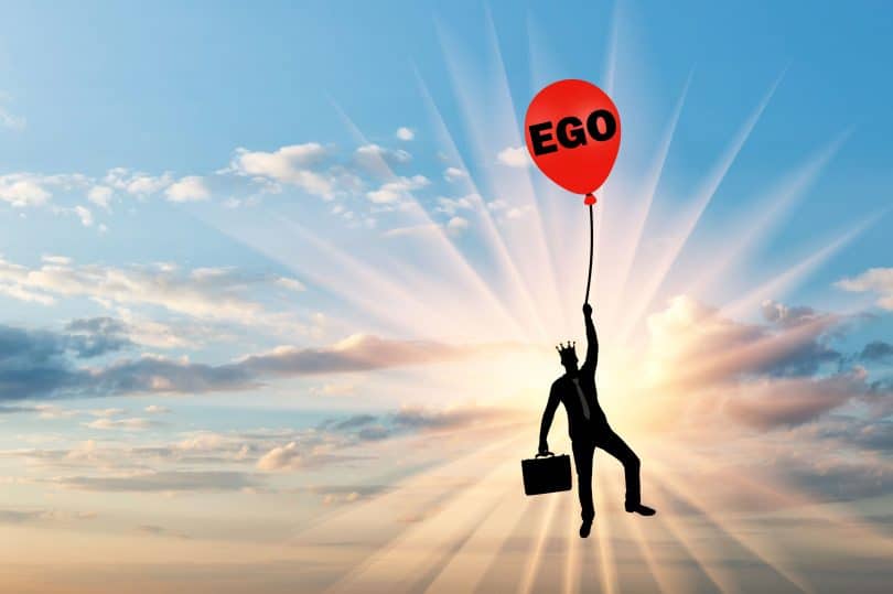 Homem carregando mala e usando uma coroa enquanto segura um balão vermelho com a a palavra "ego" desenhada.