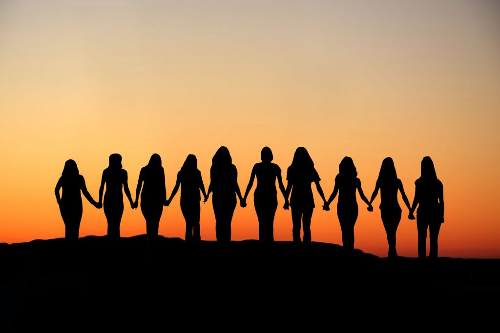 Silhueta de diversas mulheres de mãos dadas observando o pôr do sol.