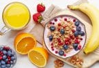 Mesa de café da manhã com uma tigela de frutas, frutas frescas cortadas e um copo de suco de laranja.
