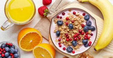 Mesa de café da manhã com uma tigela de frutas, frutas frescas cortadas e um copo de suco de laranja.