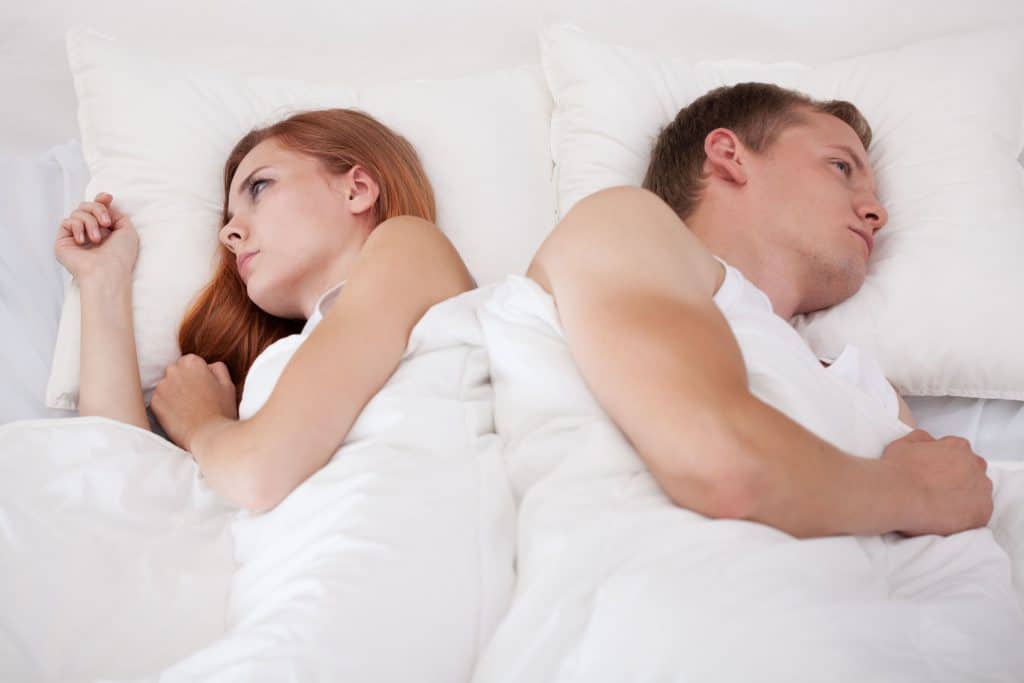casal de homem e mulher, deitados em uma cama branca, um de costas pro outro, ambos com cara de frustado.