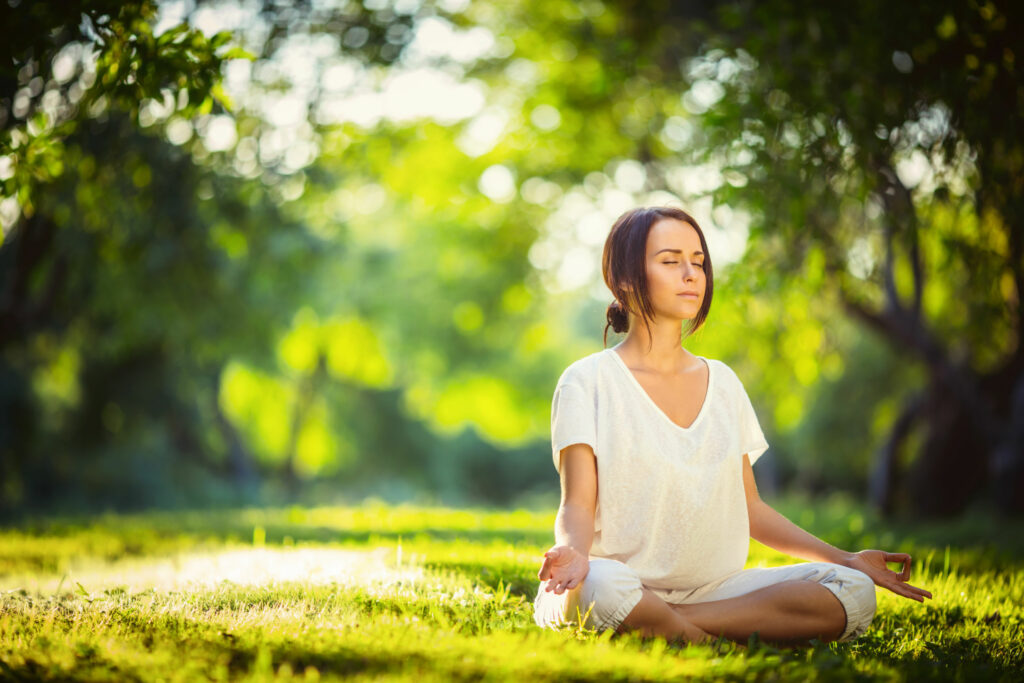 Mulher branca, vestida de branco, sentada na floresta, com as pernas cruzadas, meditando.