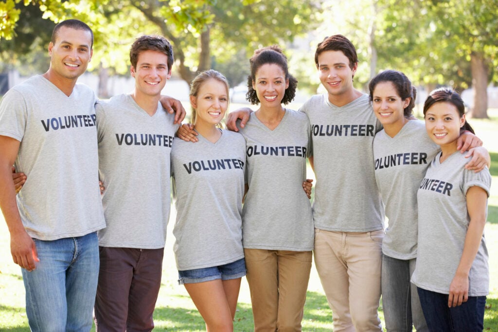 Grupo de pessoas de diversas etnias, todos sorrindo e vestindo camisetas cinzas, com a palavra voluntário escrito.