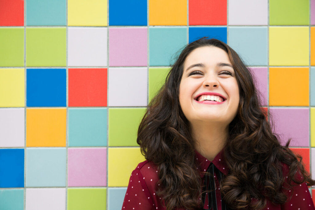 Mulher branca, jovem, sorridente, encostada em uma parede com azulejos coloridos.