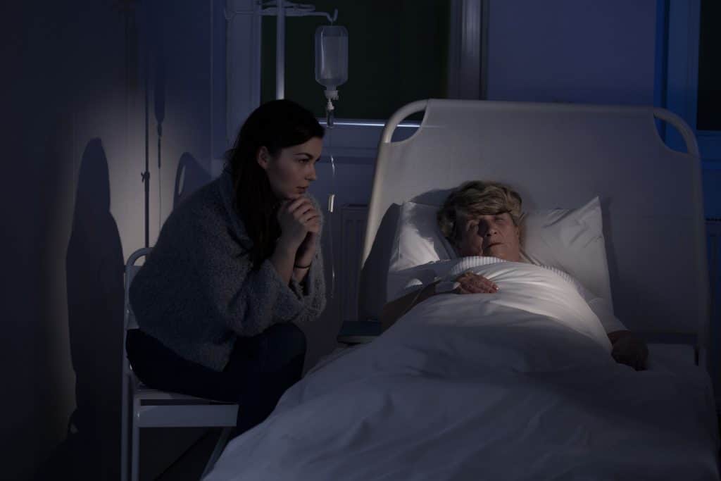 Mulher sentada ao lado de cama de hospital, observando senhora enquanto ela dorme.