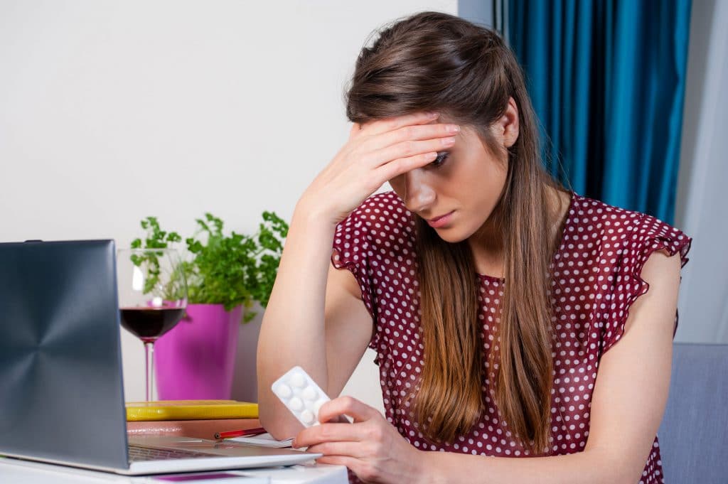 Mulher branca preocupada sentada em mesa com computador segurando cartela de pílulas anticoncepcional na sua mão.