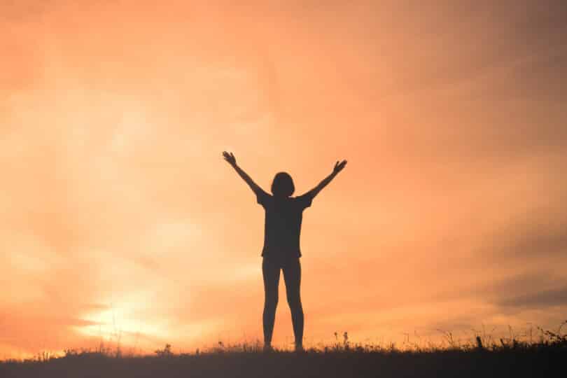 Mulher em paisagem livre ao pôr-do-sol com os braços abertos para o céu.