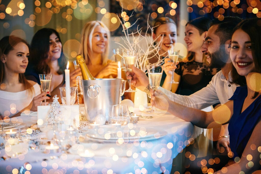 Pessoas de diversas etnias sentados em uma mesa branca, decorada para festa com coisas douradas. Cada pessoa segura uma taça de champanhe para brindar.