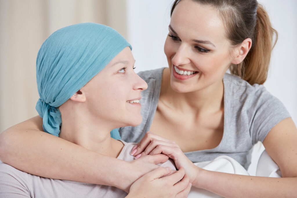 Mulher sorridente abraçando por trás a sua mãe que tem câncer e usa um lenço na cabeça, mas também sorri. 