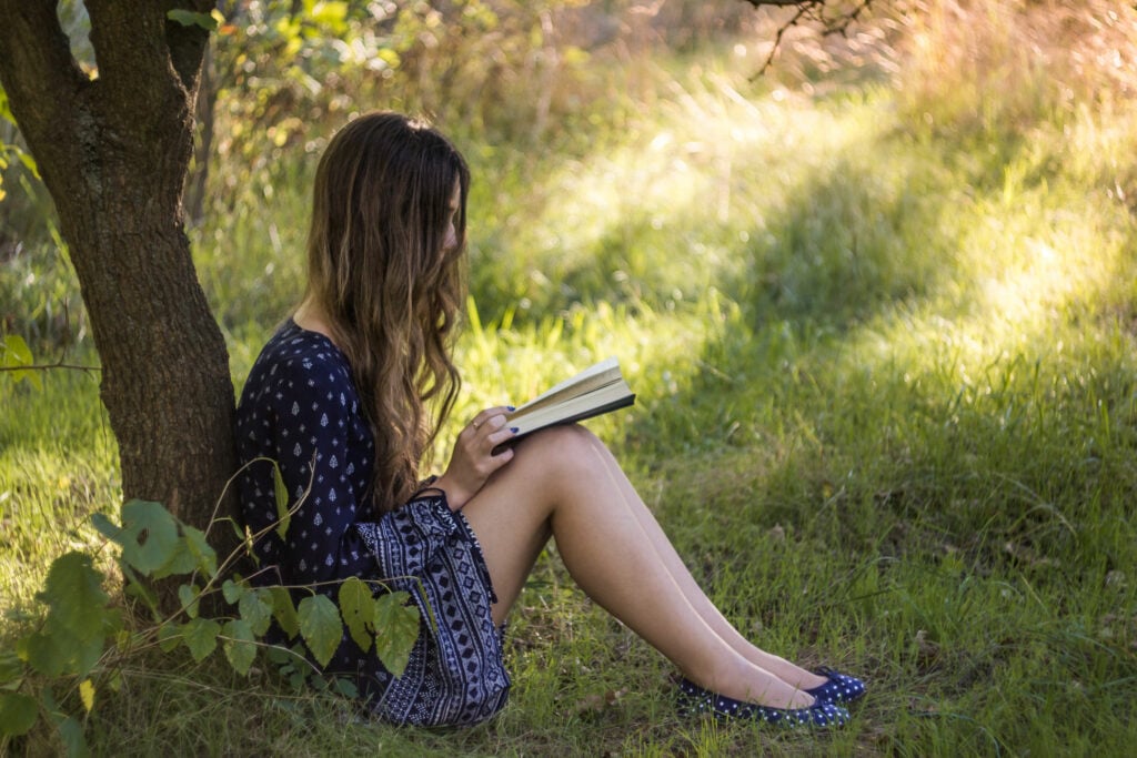 Mulher sentada embaixo de uma árvore, lendo um livro.