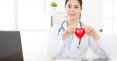 Médica em consultório usando um jaleco branco e um estetoscópio segurando um coração nas mãos.