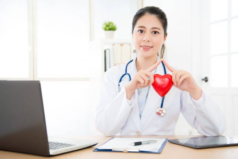Médica em consultório usando um jaleco branco e um estetoscópio segurando um coração nas mãos.