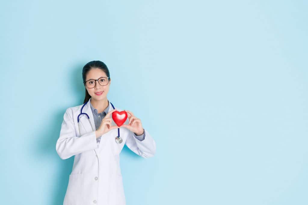 Médica com jaleco em fundo azul segurando um coração vermelho.