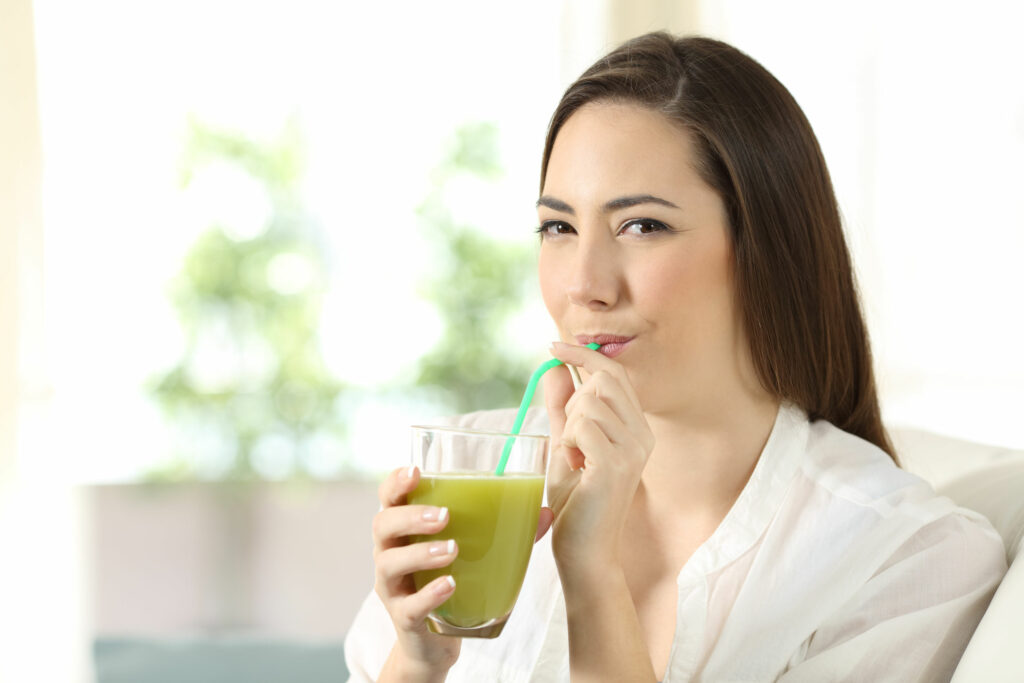 Jovem tomando suco verde enquanto está sentada no sofá.