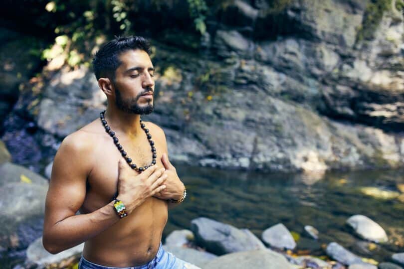 Homem meditando com as mãos sobre o peito nu. Ele está sobre as rochas que permeiam um riacho.