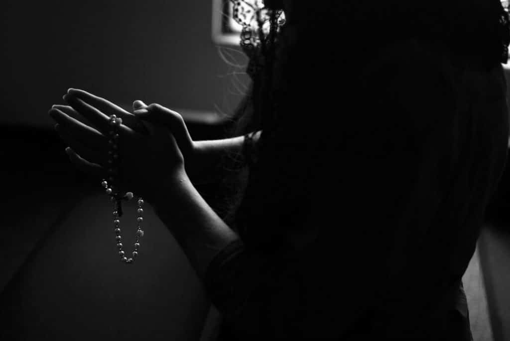 Foto preta e branco de mulher orando, segurando um terço nas suas mãos.