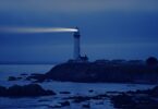 Farol emitindo luz para o mar durante a noite, auxiliando no caminho dos navegantes. Ele está sobre uma elevação de terra, bem como as casas ao redor.