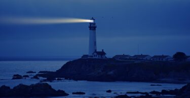 Farol emitindo luz para o mar durante a noite, auxiliando no caminho dos navegantes. Ele está sobre uma elevação de terra, bem como as casas ao redor.