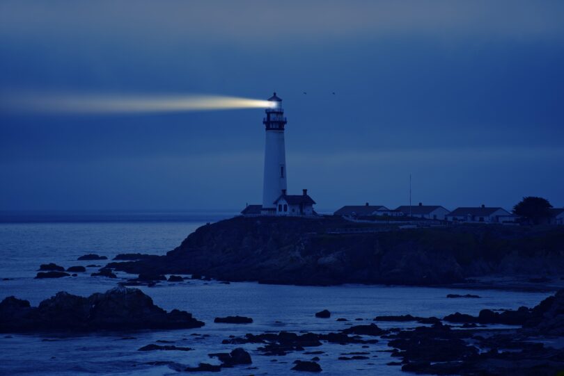 Farol emitindo luz para o mar durante a noite, auxiliando no caminho dos navegantes. Ele está sobre uma elevação de terra, bem como as casas ao redor.