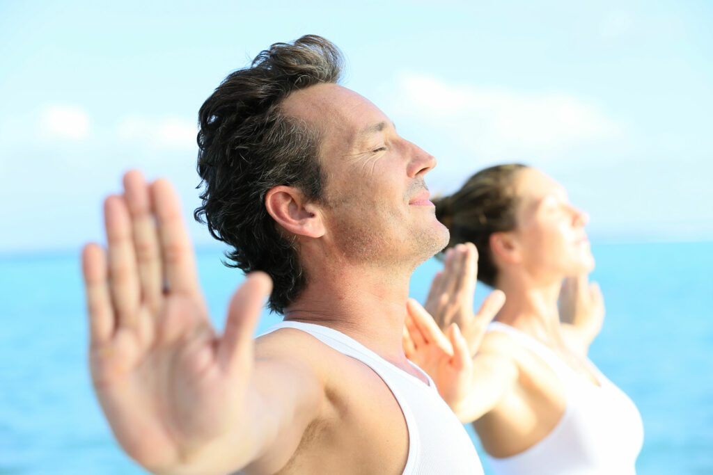 Homem e mulher vestidos de branco, praticando yoga, com visão do mar ao fundo.