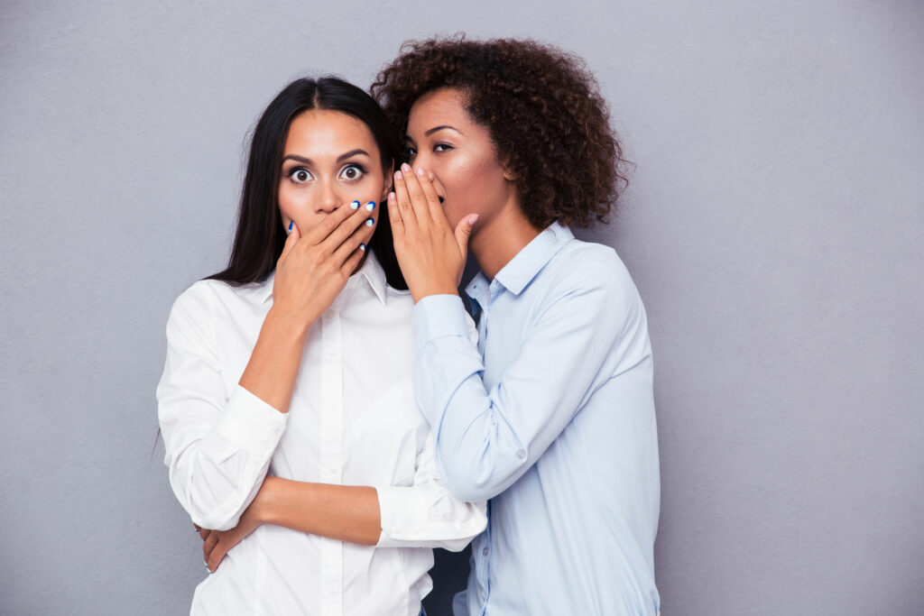 Duas mulheres negras, jovens, uma contando um segredo no ouvido da outra, que mostra surpresa.