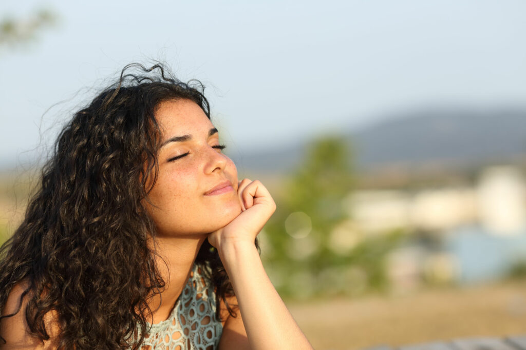 Mulher branca, jovem, sentada no chão, tomando sol no rosto, com os olhos fechados.