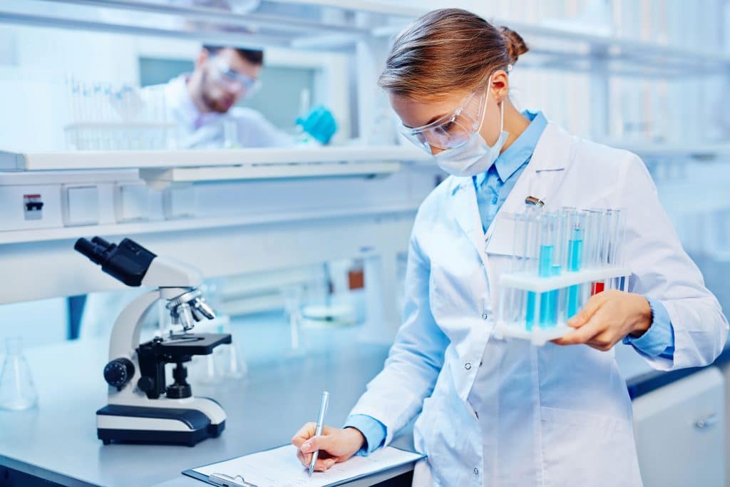 Mulher em laboratório científico, fazendo anotações em uma prancheta enquanto segura com suporte cheio de tubos de ensaio com líquido azul.