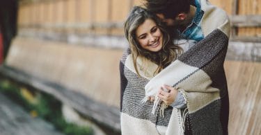 Casal enrolado em um cobertor no foco com mulher sorrindo e homem com cabeça apoiada no cabelo dela